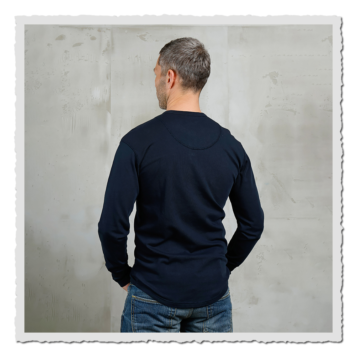 Rückansicht eines Models im dunkelblauen Langarmshirt vor heller, leicht strukturierter Wand.